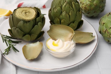 Delicious cooked artichokes with tasty sauce served on white tiled table, closeup