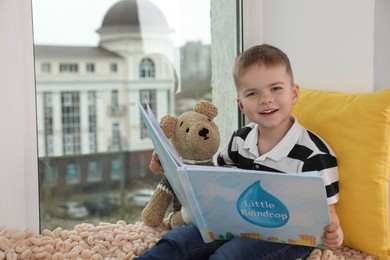 Photo of Cute little boy with toy bear reading book near window at home, space for text