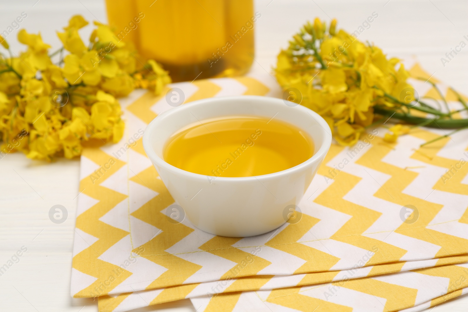 Photo of Rapeseed oil in bowl and beautiful yellow flowers on white table, closeup