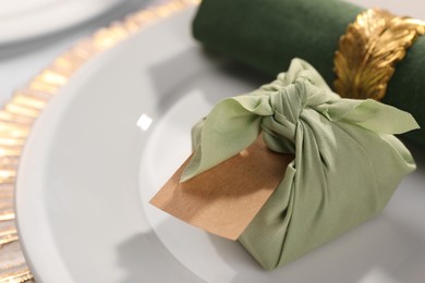 Photo of Furoshiki technique. Gift packed in green fabric, blank card and napkin on plate, closeup. Space for text