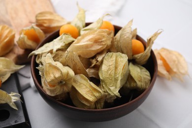 Ripe physalis fruits with calyxes in bowl on white tiled table, closeup