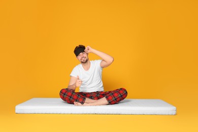 Photo of Smiling man in sleeping mask sitting on soft mattress and showing thumb up against orange background