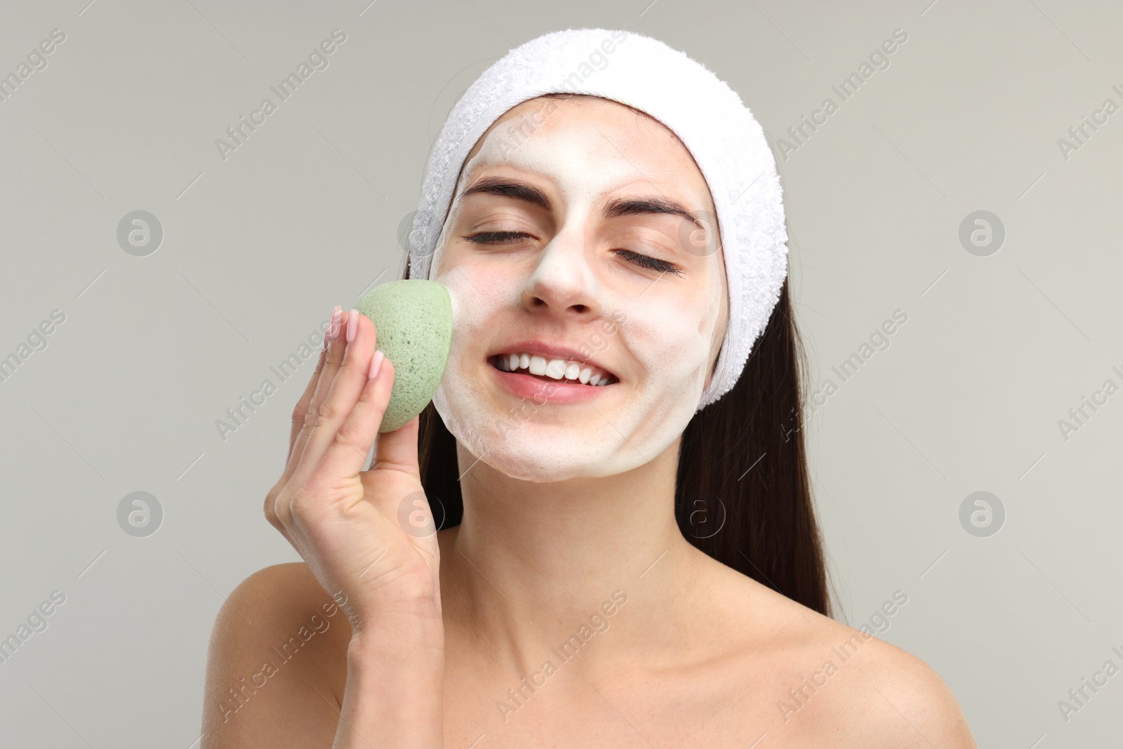 Photo of Young woman with headband washing her face using sponge on light grey background