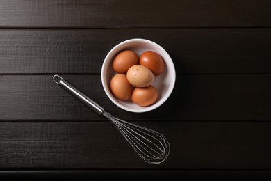 Metal whisk and raw eggs in bowl on dark wooden table, top view