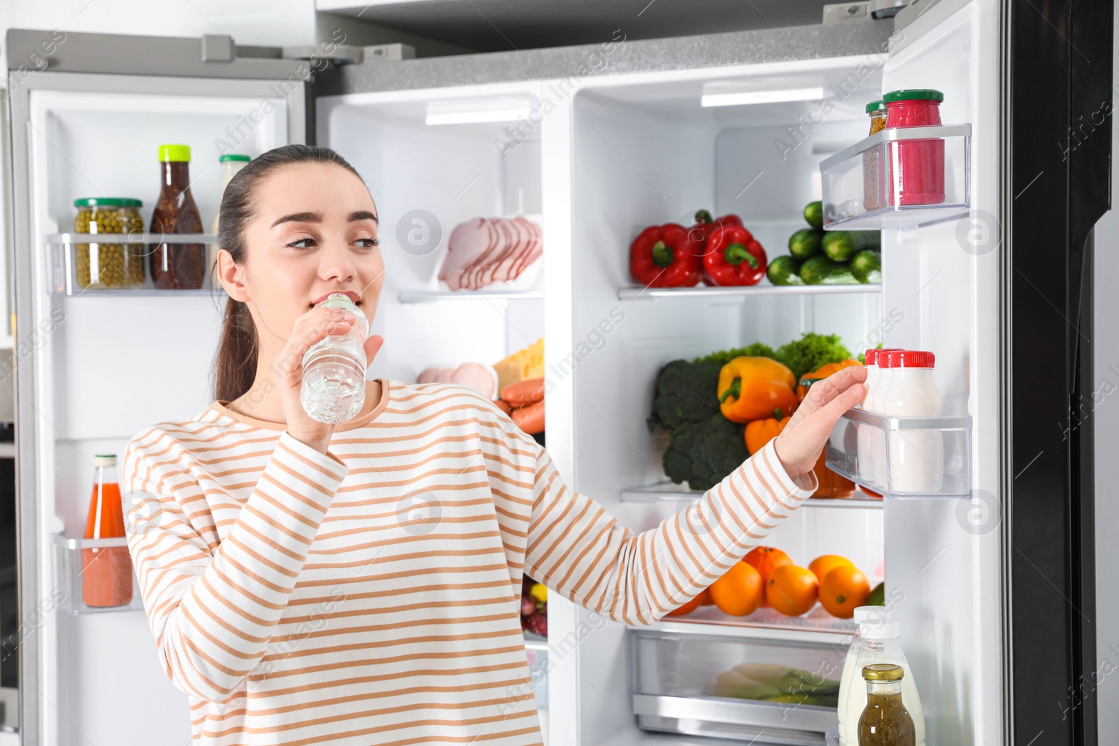 Photo of Young woman drinking water near open refrigerator indoors