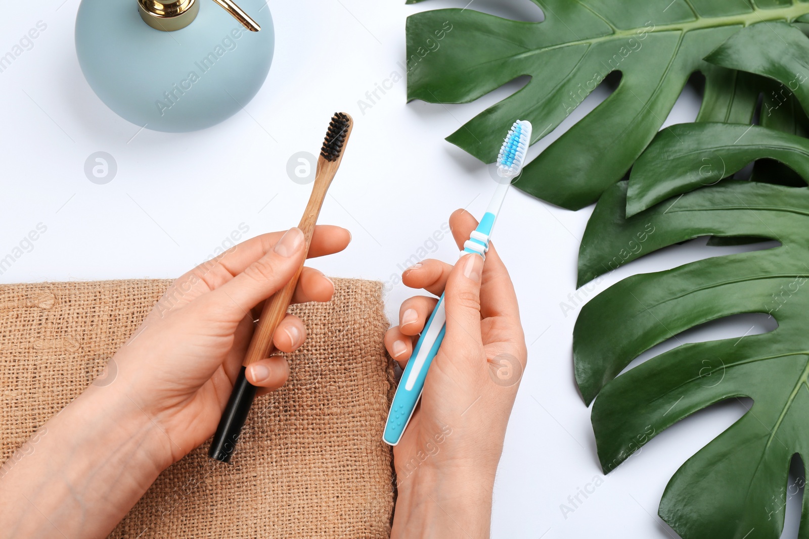 Photo of Woman holding natural bamboo and plastic toothbrushes over white table, above view