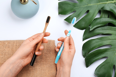 Woman holding natural bamboo and plastic toothbrushes over white table, above view