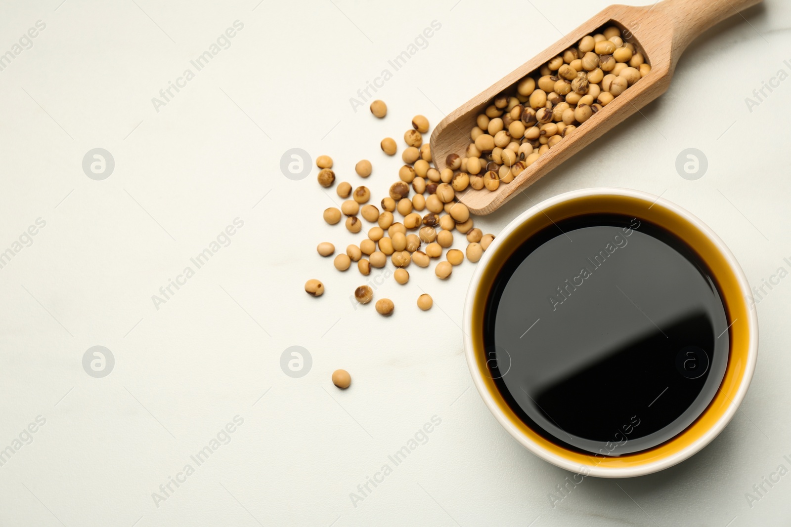 Photo of Soy sauce in bowl and scoop with soybeans on white table, flat lay. Space for text