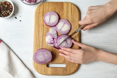 Photo of Young woman cutting red onion on wooden board at white table, top view