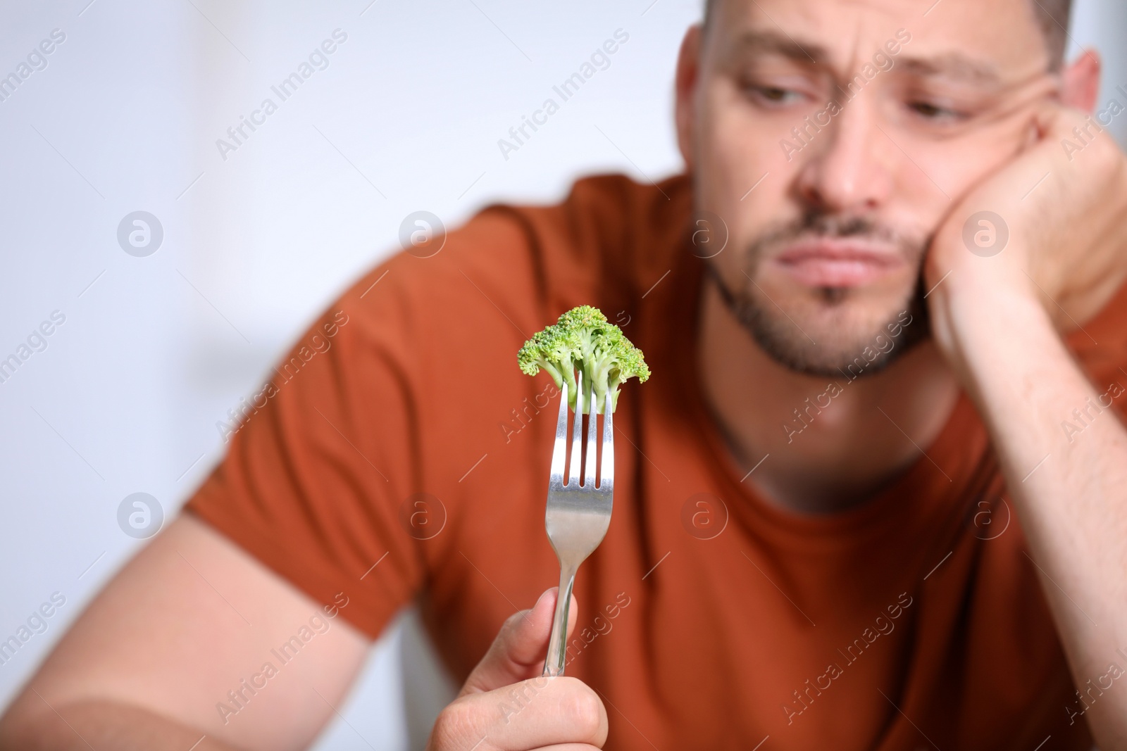 Photo of Unhappy man with broccoli on fork against light background, closeup