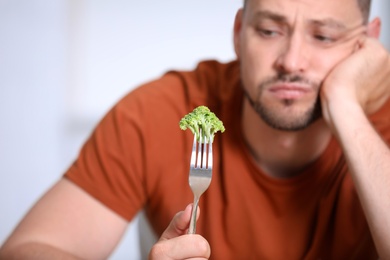 Unhappy man with broccoli on fork against light background, closeup