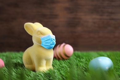 Image of COVID-19 pandemic. Easter bunny in protective mask and painted eggs on green grass, space for text