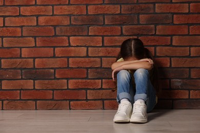 Photo of Child abuse. Upset little girl sitting on floor near brick wall indoors, space for text