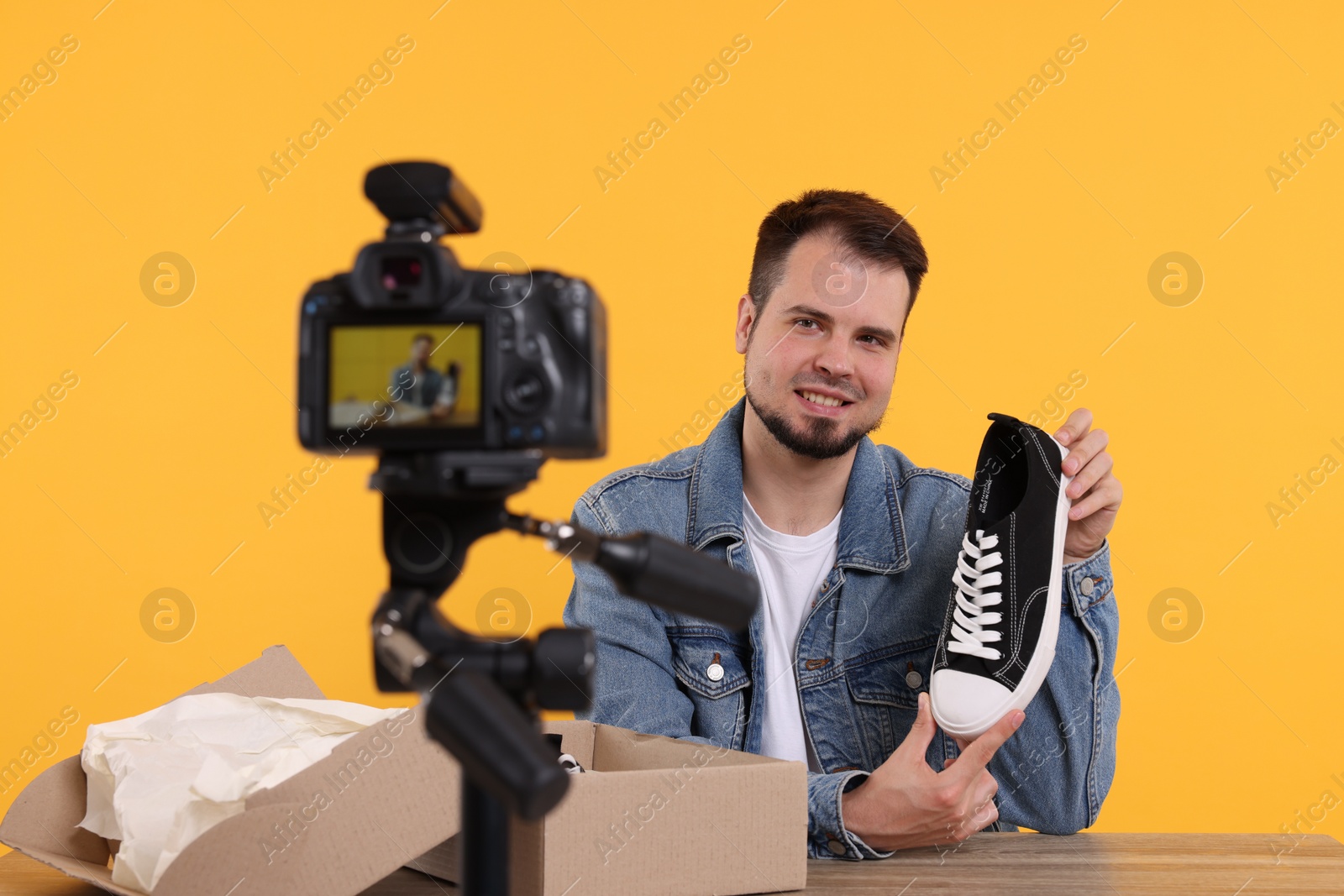 Photo of Smiling fashion blogger showing sneaker while recording video at table against orange background