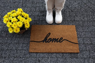 Photo of Woman in stylish boots near doormat with word Home and flowers on pavement, closeup