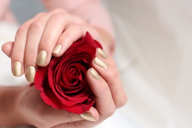 Woman with gold manicure holding rose on blurred background, closeup. Nail polish trends