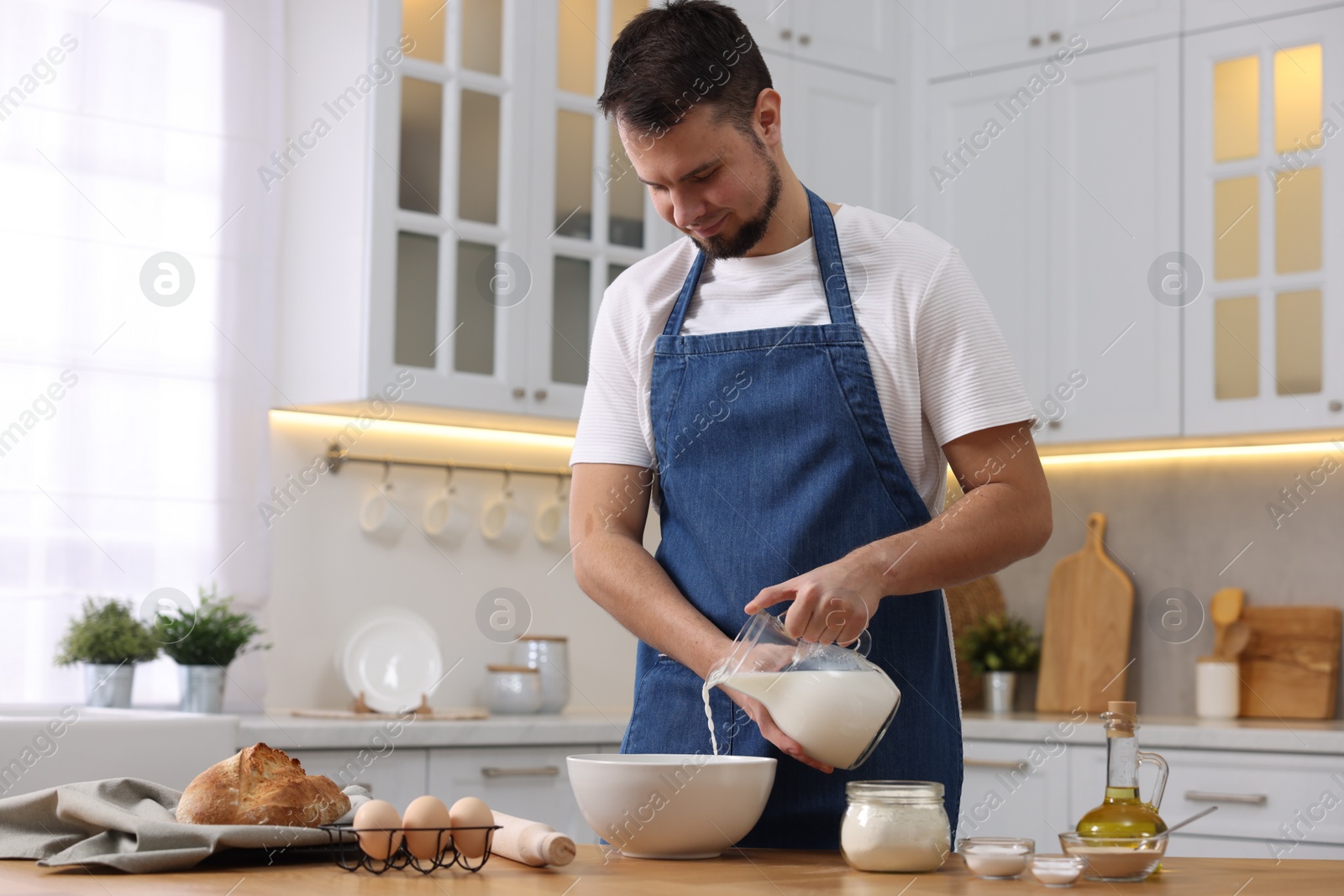 Photo of Making bread. Man pouring milk into bowl at wooden table in kitchen