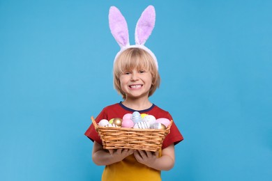 Photo of Happy boy in bunny ears headband holding wicker basket with painted Easter eggs on turquoise background