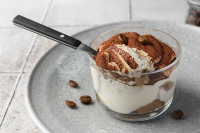 Photo of Delicious tiramisu in glass, coffee beans and spoon on light tiled table, closeup