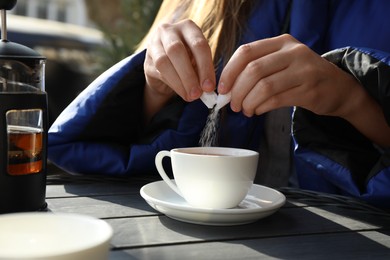 Woman adding sugar into cup of tea at black wooden table in outdoor cafe, closeup