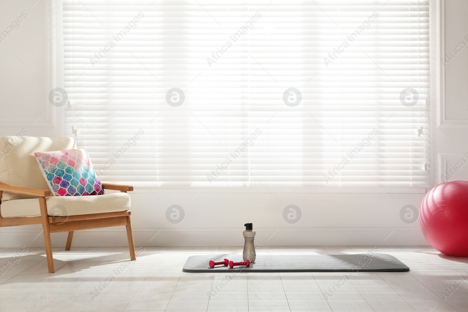 Photo of Exercise mat, dumbbells, bottle and fitness ball near armchair in spacious room