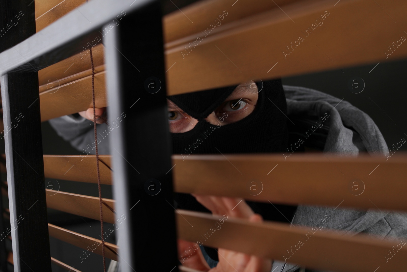 Photo of Masked man spying through window blinds indoors. Criminal offence