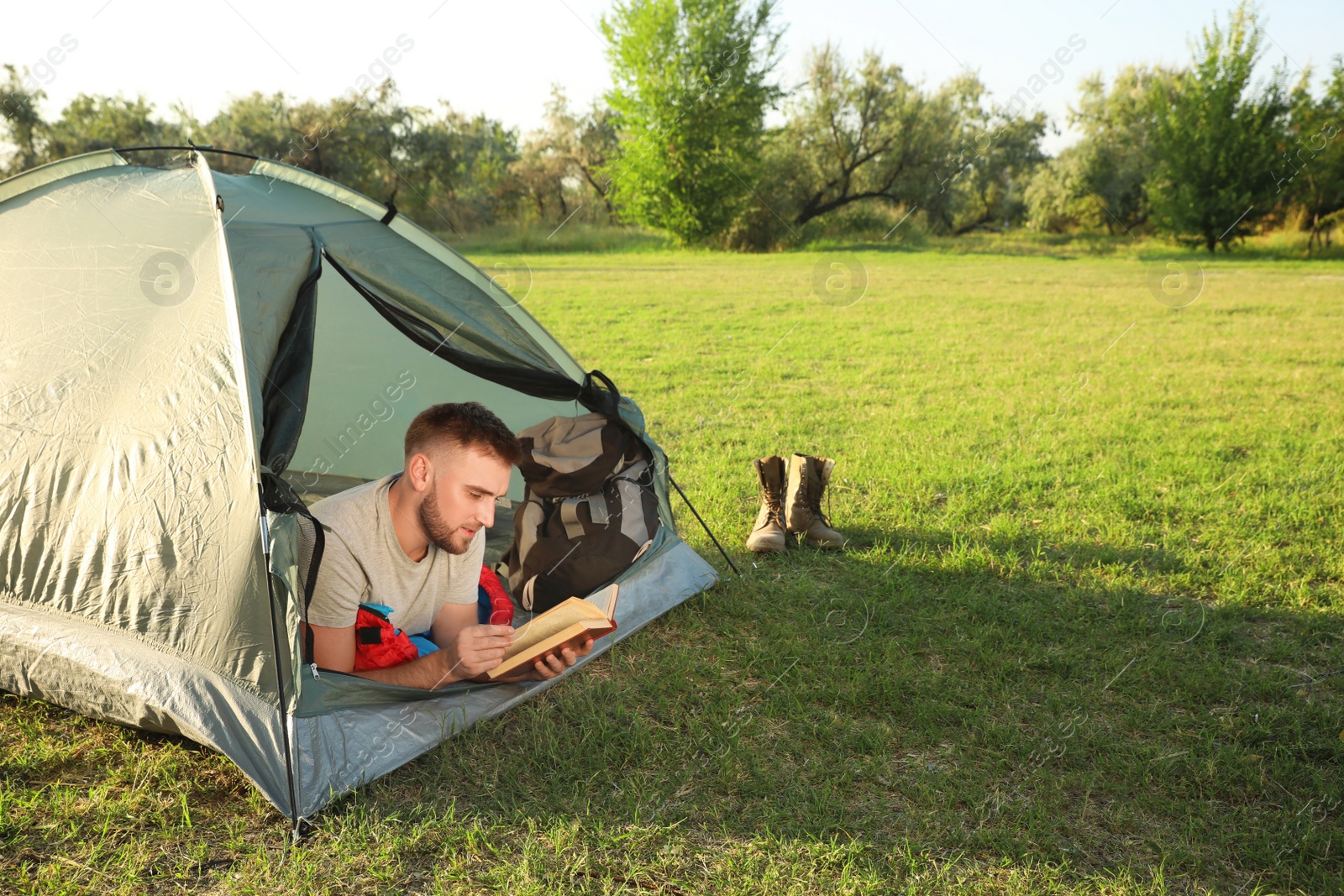 Photo of Young man in sleeping bag with book lying inside camping tent