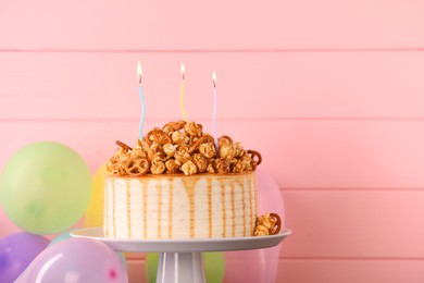 Photo of Caramel drip cake decorated with popcorn and pretzels near balloons against pink wooden background, space for text