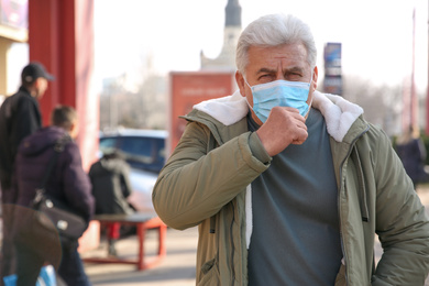 Photo of Senior man with medical mask coughing on city street. Virus protection