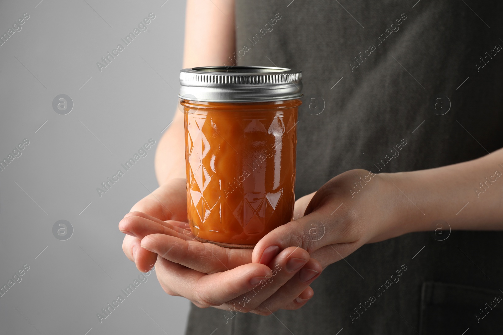 Photo of Woman holding glass jar of delicious persimmon jam on gray background, closeup. Space for text