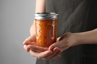 Photo of Woman holding glass jar of delicious persimmon jam on gray background, closeup. Space for text