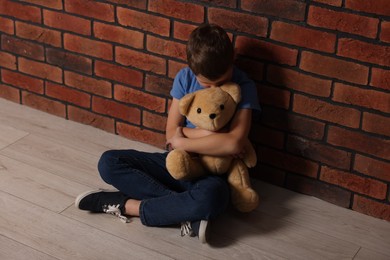 Child abuse. Upset boy with teddy bear sitting on floor near brick wall indoors