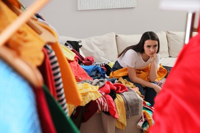 Upset young woman with lots of clothes on sofa in room. Fast fashion