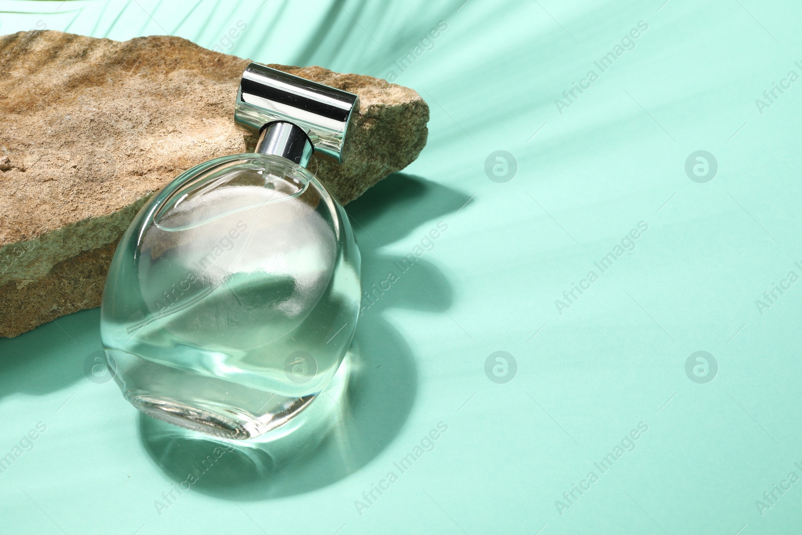 Photo of Bottle of luxury perfume in sunlight and stone on turquoise background, space for text