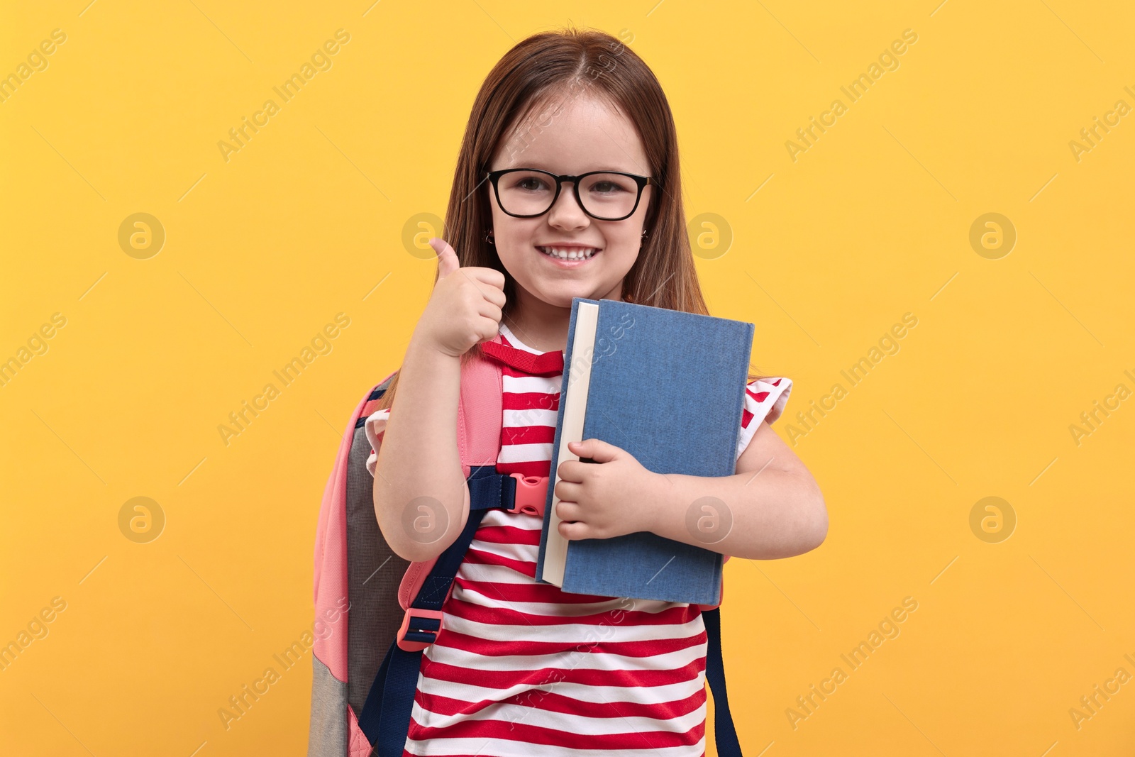 Photo of Cute little girl with backpack and book showing thumbs up against orange background