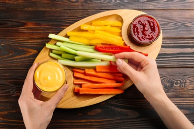 Woman holding sauce and celery stick above tray with different vegetables at wooden table, top view