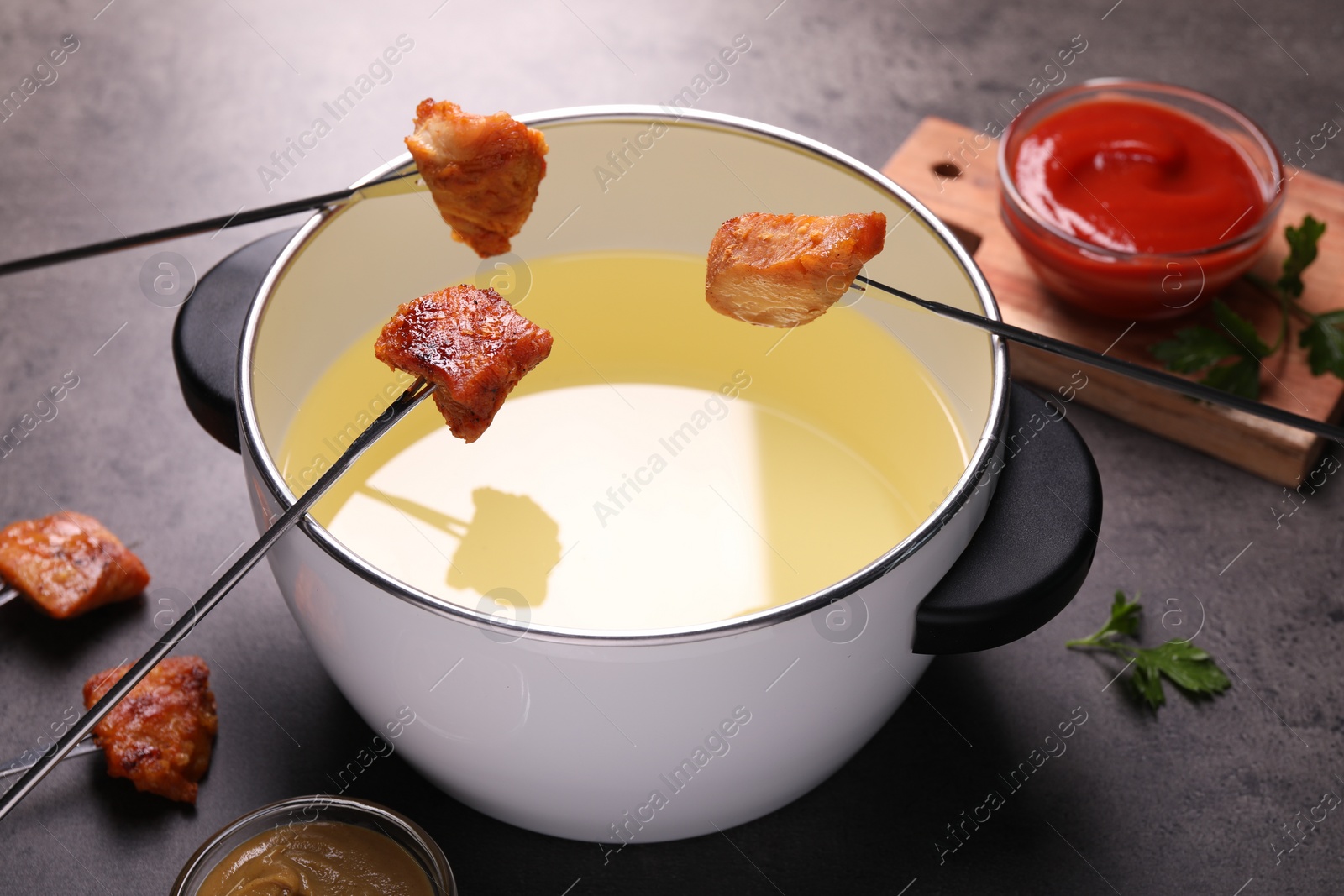 Photo of Oil in fondue pot, forks with fried meat pieces and sauces on grey table, closeup