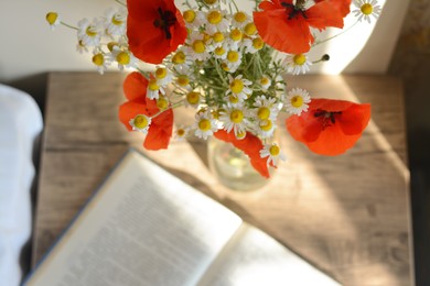 Beautiful bouquet of poppies and chamomiles near open book indoors, focus on flowers