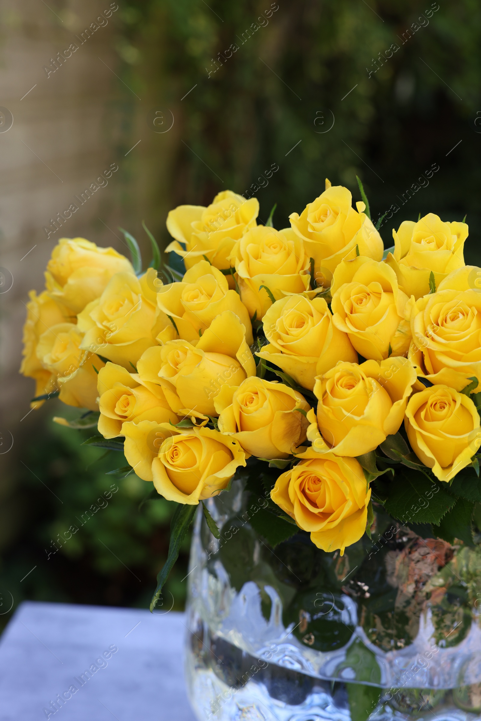 Photo of Beautiful bouquet of yellow roses in glass vase on light table outdoors