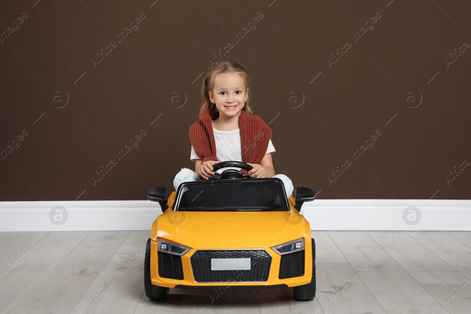 Photo of Cute little girl driving children's electric toy car near brown wall indoors