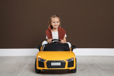 Cute little girl driving children's electric toy car near brown wall indoors