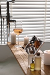 Photo of Wooden tray with tablet, wine, candles and toiletries on bathtub in bathroom