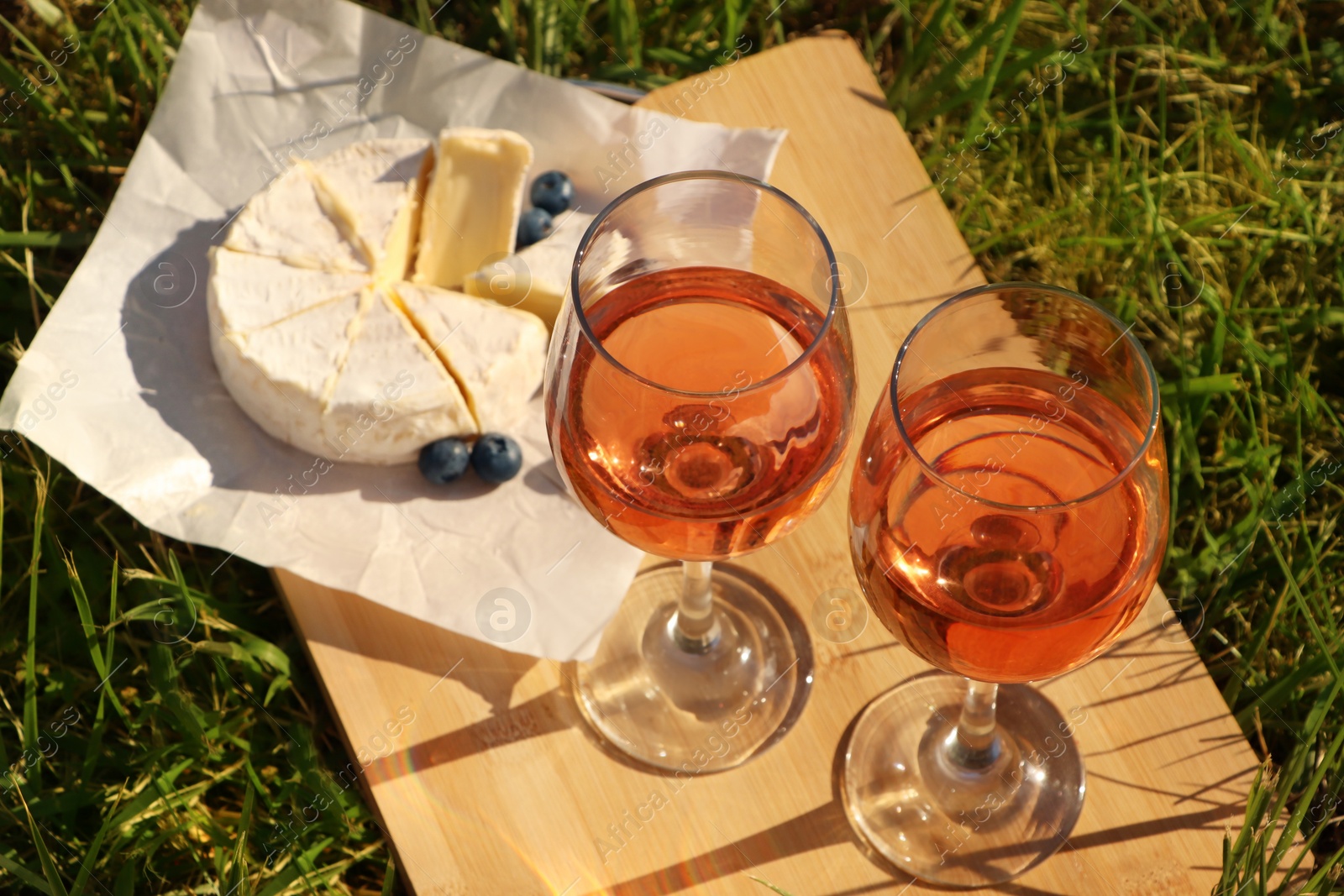 Photo of Wooden board with glasses of delicious rose wine, cheese and blueberries on green grass outdoors