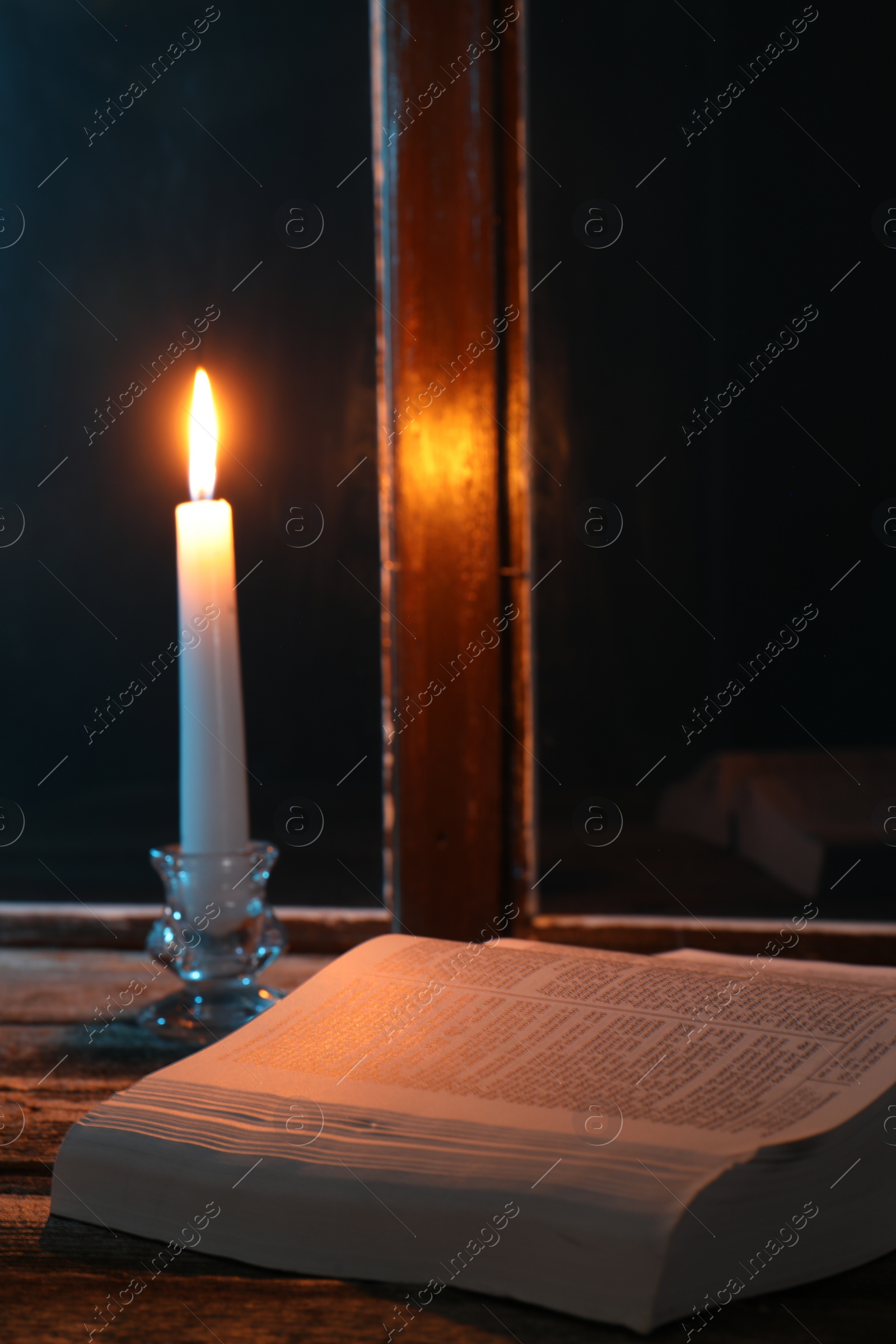 Photo of Burning candle and Bible on wooden table near window at night