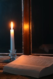 Photo of Burning candle and Bible on wooden table near window at night