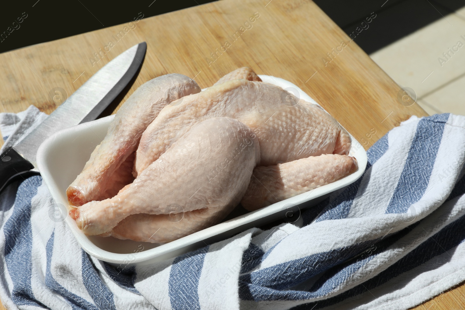 Photo of Fresh raw whole chicken in plastic container, knife and napkin on wooden table