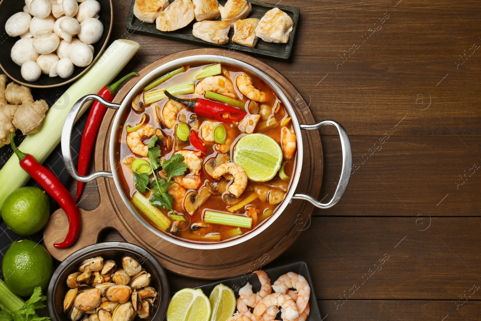 Photo of Saucepan with delicious Tom Yum soup and ingredients on wooden table, flat lay. Space for text