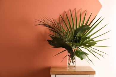 Photo of Fresh leaves of tropical plants in vase on table near color wall. Home decoration