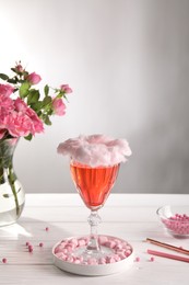 Photo of Cotton candy cocktail in glass, marshmallows and vase with pink roses on white wooden table against gray background