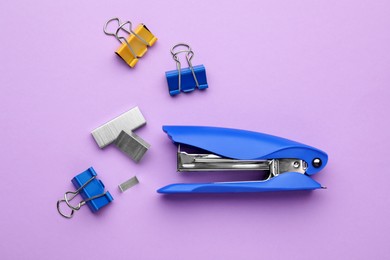 Photo of New bright stapler with staples and blinder clips on violet background, flat lay. School stationery